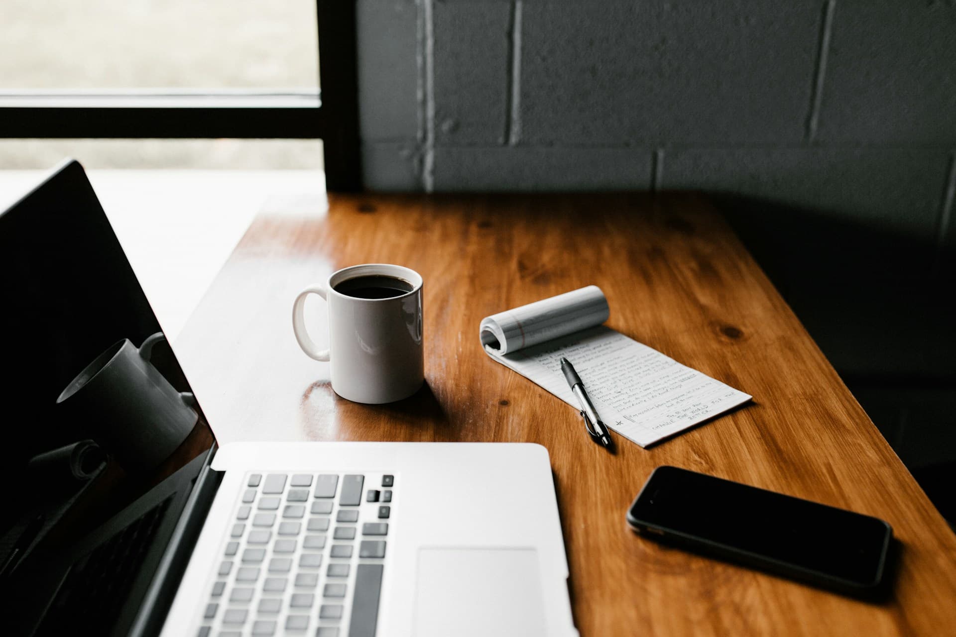 A wooden desk with a laptop, coffee, notepad and a phone.